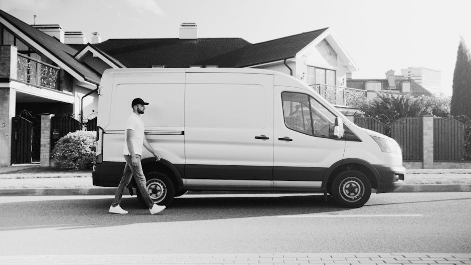 A person dressed in full protective clothing, including a white disposable coverall, hood, face shield, gloves, and shoe covers, stands outdoors next to a white van with its sliding side door open, du