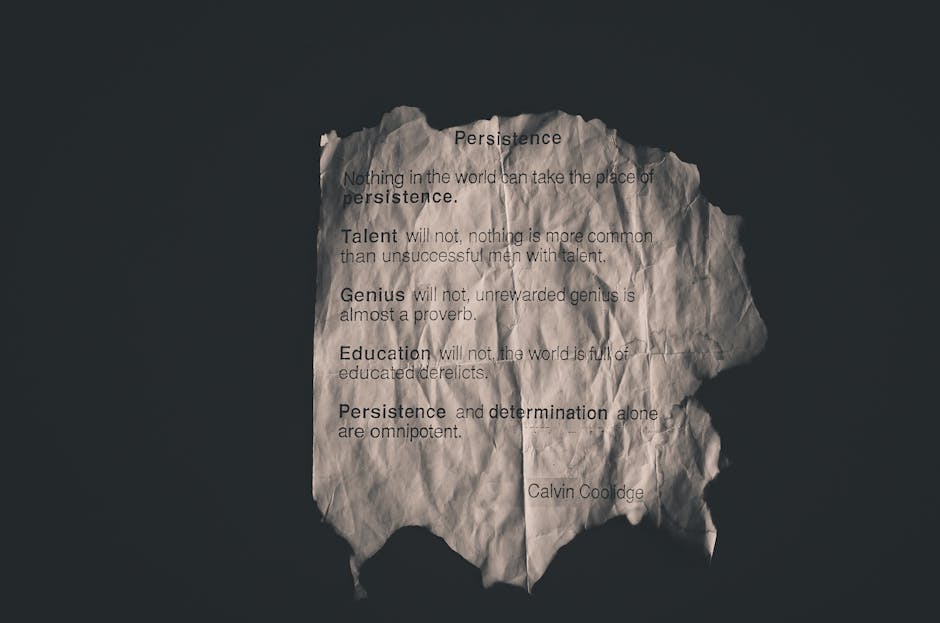 A crumpled piece of white paper with torn edges is photographed against a dark background. The paper features printed text displaying motivational quotes about persistence, talent, genius, and education, attributed to Calvin Coolidge. The lighting highlights the creases and irregular shape of the paper, emphasizing its textured surface. The message on the paper contains various lines of text, some with slight distortions due to the crumpling, and the shadows cast by the paper add depth to the image. The scene appears to be closely focused, with the paper occupying most of the frame, creating a stark contrast between the white paper and the dark backdrop. This image visually emphasizes the concept of perseverance and resilience, often associated with themes of home relocation and moving, which aligns subtly with services like those offered by Man with Van Old Malden, especially in sensitive discussions around hidden fees or confusing quotes in property moves.