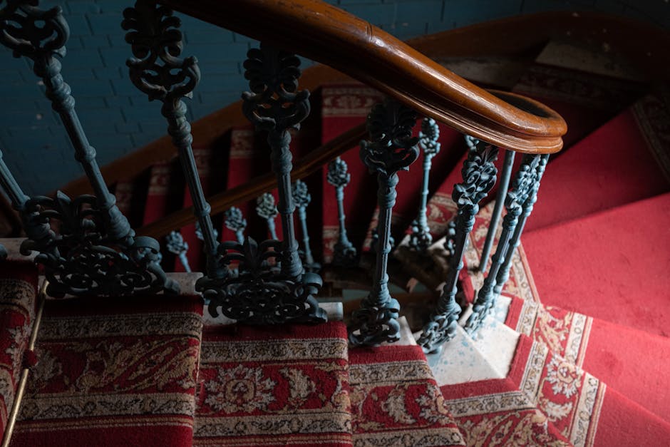 A view from the top of a narrow Victorian staircase inside a property, showing red patterned carpeted steps with a decorative, dark metal banister featuring intricate scrollwork. The staircase curves downward, with a wooden handrail at the top. The area is lit with soft, natural light, highlighting the textured carpet and the detailed ironwork of the balustrade. This image depicts the challenging environment faced during home relocation or furniture transport in Old Malden, with [COMPANY_NAME] providing professional moving services that include navigating tight staircases and ensuring safe packing and loading processes for residents and their belongings.