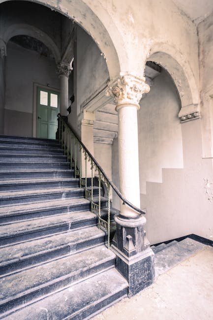 Inside an old Victorian building featuring a narrow staircase made of dark, polished stone with a black and gold metal railing on the left side. The staircase ascends toward a green door visible at the top, set within an arched doorway. The surrounding walls and ceiling have a worn, aged appearance with peeling paint and exposed plaster, highlighting the historic character of the interior. To the right of the staircase, there is a partially visible curved white plaster wall with decorative columns supporting the archways, creating a spacious, open entrance area. The lighting is soft and natural, coming from an unseen source, illuminating the textured surfaces and the architectural details of the entryway. This setting depicts a typical phase of a home relocation or moving process within an older property, where careful handling of furniture and materials might be necessary to navigate the narrow stairs, as seen by the pristine condition of the staircase and the historical features of the space. Man with Van Old Malden may be involved in such residential removals, planning transport and packing strategies suitable for Victorian staircases and compact interiors.