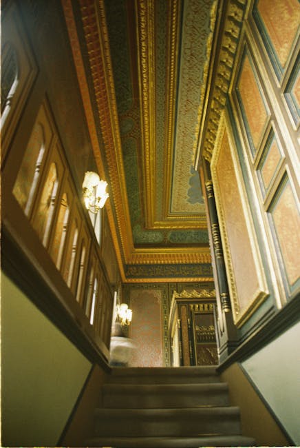 A view from the top of a narrow Victorian staircase inside a property, showing red patterned carpeted steps with a decorative, dark metal banister featuring intricate scrollwork. The staircase curves downward, with a wooden handrail at the top. The area is lit with soft, natural light, highlighting the textured carpet and the detailed ironwork of the balustrade. This image depicts the challenging environment faced during home relocation or furniture transport in Old Malden, with [COMPANY_NAME] providing professional moving services that include navigating tight staircases and ensuring safe packing and loading processes for residents and their belongings.