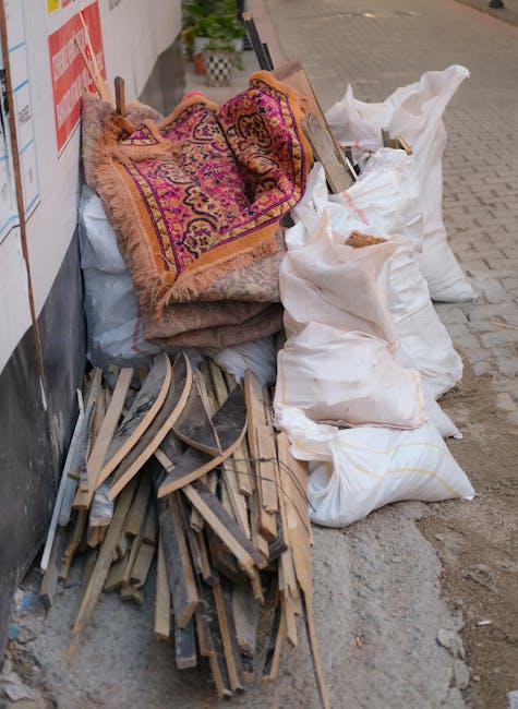 A pile of various household items and waste materials placed outside a building for clearance or disposal. The collection includes a rolled-up, ornate, multicolored rug with fringed edges, several large white fabric sacks filled with unknown contents, and a disorganized stack of wooden furniture parts or frames. The items are situated on a paved pavement area next to a building wall, with some of the furniture parts leaning against the wall and others lying on the ground. The scene captures the typical appearance of items prepared for a home relocation or clearance service, where Man with Van Old Malden assists with furniture transport and waste removal. The overall setting suggests an ongoing clearance process, possibly related to house removals or renovations, with the loading process awaiting collection by waste disposal or recycling services.