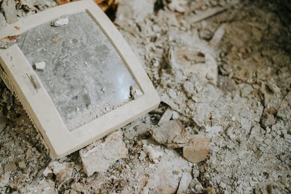 A dirty, dust-covered old wall-mounted light switch with a plastic cover lying on a rough, uneven surface composed of loose dirt, small rocks, and debris. The switch appears to be disconnected from its wiring and is surrounded by scattered fragments of concrete, brick, and dust, indicating ongoing building or renovation work. In the background, the textured surface continues with more dirt and rubble, suggesting an unfinished or dismantled interior space. This scene reflects a typical environment encountered during house clearance or refurbishment, where removal of fixtures like light switches is part of the packing and home relocation process, as handled by Man with Van Old Malden. The lighting is natural, highlighting the coarse material textures and the state of disassembly, matching the context of preparing a property for clearance or move.