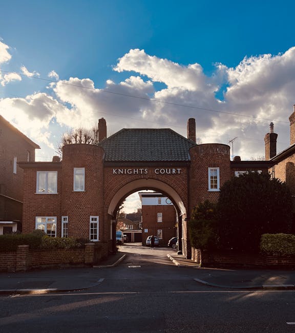 Photograph of the entrance to Knights Court, an archway flanked by brick residential buildings with white-framed windows, situated in Old Malden. The archway leads to a residential street with parked cars visible in the background. The sky above features clouds illuminated by sunlight, creating a partly cloudy scene. To the right of the entrance, there is a bush, and on the left, parts of neighboring buildings are visible. The scene is well-lit with natural daylight, capturing the typical urban surroundings of a local housing area, which is relevant to home relocation and furniture transport logistics as seen in professional removals by Man with Van Old Malden.