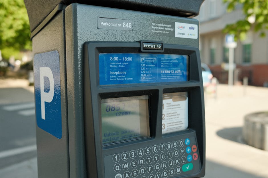Close-up view of a parking meter situated on a residential street with trees and houses visible in the background. The meter has a blue 'P' sign on the side and a blue digital display showing parking charges and instructions in Polish, including rates and timings. The payment keypad, with numbered buttons and function keys, is positioned below the display. The surrounding area appears bright with natural daylight, suggesting a daytime setting. This parking meter is part of the parking arrangements that Kingston Council manages, relevant to home relocation and furniture transport in the area. Man with Van Old Malden utilises such parking facilities during their professional removals and moving services, ensuring compliant and efficient logistics for house removals in Kingston and Old Malden.