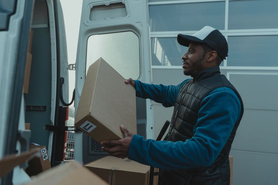 A man wearing a grey cap, black padded vest, and blue long-sleeved shirt is seen loading a medium-sized cardboard box into the back of a white moving van. The van's interior is visible, showing severa