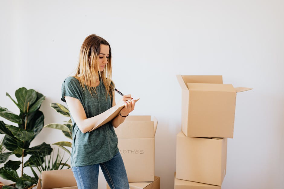 Inside a residential property, a young woman with light brown hair is standing among several cardboard moving boxes, some open and others closed, with handwritten labels such as 'clothes.' She is wearing a casual grey t-shirt and blue jeans, and is taking notes on a notepad with a pen, likely preparing a house moving checklist. Behind her, there is a large green houseplant with broad leaves, positioned against a plain white wall, contributing to a bright and neutral indoor environment. To her right, a stack of open cardboard boxes is visible, ready for packing or organizing personal belongings, supporting the process of home relocation. The scene captures a moment of planning and packing, with natural indoor lighting illuminating her and the boxes, illustrating the logistics involved in furniture transport and packing for a household move, as part of a professional removals service like Man with Van Old Malden.
