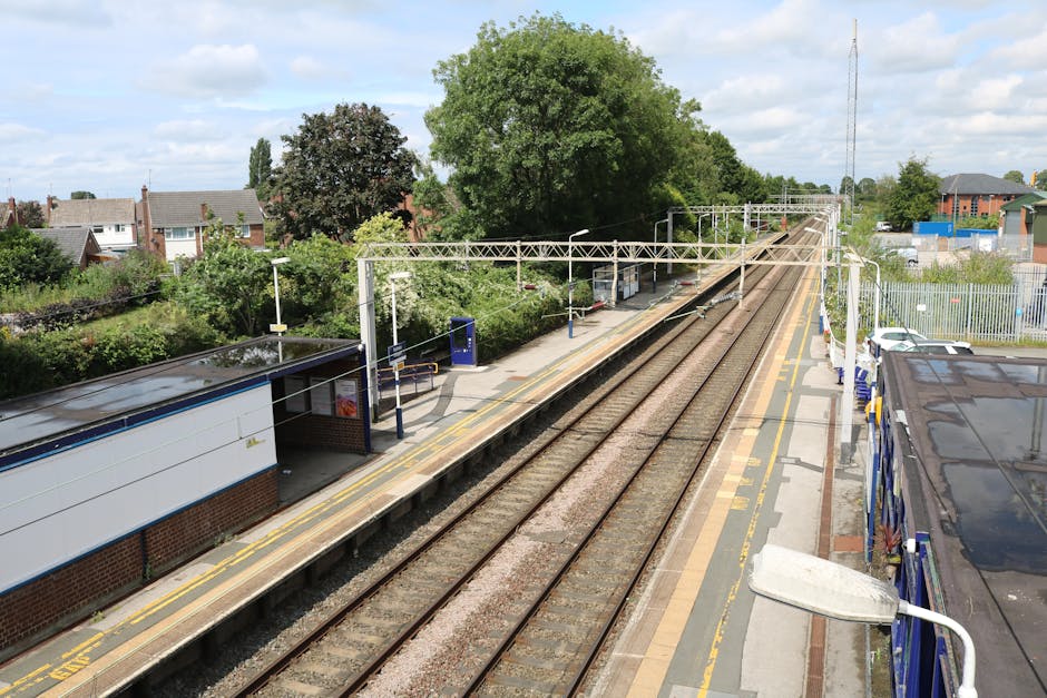 An elevated view of a small train station platform adjacent to railway tracks during daytime. The platform features a sheltered waiting area with a brick wall, glass windows, and a seating area, along with various signage and lighting fixtures. Several cardboard boxes and wrapped furniture items are positioned along the edge, ready for loading onto a nearby blue van parked on the right side of the image. The van is partially visible, with its roof and side doors in view, indicating a home relocation process managed by Man with Van Old Malden. The surrounding environment includes lush green trees, residential houses with pitched roofs, and a tall communication mast in the distance, all under a partly cloudy sky. This setting illustrates the logistics involved in local moving and furniture transport, emphasizing efficient loading in an outdoor setting as part of house removals.