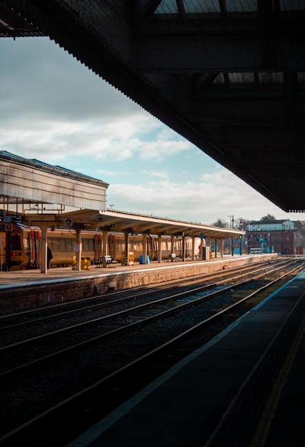 A view of a train station platform at Malden Manor with train tracks running alongside. The platform features a sheltered waiting area with multiple benches and a canopy extending over part of it. The sky above is partly cloudy, with some sunlight illuminating the scene. In the background, there are station buildings and other infrastructure. The image captures the environment where people may load or unload belongings during home relocations, fitting with the context of house removals and furniture transport for local moving services, as provided by Man with Van Old Malden.