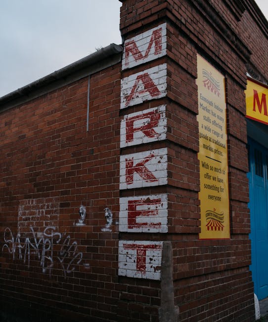 Close-up view of a brick building corner with a vertically painted sign spelling 'MARKET' in red letters on a white background on the wall. To the right, a yellow informational poster about a market is affixed next to a blue garage door, partially visible. In the lower left corner, grey graffiti tags are painted on the brickwork. The scene is illuminated with natural light, revealing weathered brick textures and slight discoloration on the sign. This setting depicts an exterior urban environment likely associated with local shopping or market activities, relevant to house and shop removals, and moving logistics by Man with Van Old Malden.