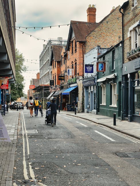 A daytime street scene on Old Malden High Street featuring a mix of commercial and residential buildings, with some structures made of red brick and others painted in shades of blue and grey. The sky is overcast, and decorative string lights are hanging across the narrow road, which is lined with small shops and eateries. Several pedestrians are walking along the pavement, including a person pushing a stroller, while others browse storefronts. On the right side, a van is parked near the curb, and a few individuals are engaged in loading or unloading furniture, boxes, and packing materials, indicating a home or business relocation process. Visible items include cardboard boxes, soft fabric wrapping, and protective blankets being used to secure furniture for transport. The scene reflects an active moving environment consistent with professional removals services, such as those offered by Man with Van Old Malden, during a furniture transport or packing and moving operation in preparation for a house or shop move on a busy high street.
