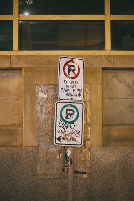 A metal pole mounted against a stone wall exterior of a building, displaying two street parking and loading restrictions signs. The upper sign indicates no parking except by permit from 7 am to 6 pm, Monday to Friday, with a red circle and slash over a 'P' symbol. The lower sign shows designated parking hours from 8 am to 6 pm, Monday to Friday, with a green circle around a 'P' symbol and a hand gesture icon, possibly indicating loading or unloading permitted during these times. Part of a window with a green-tinted glass pane and wooden framing is visible at the top. This signage is relevant for house removals or moving services, as it informs about parking and loading regulations in Old Malden, supporting the logistical planning for a home relocation by Man with Van Old Malden.