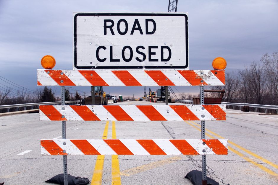 A road closure barricade positioned across an asphalt street during daytime, with a large white sign displaying 'ROAD CLOSED' in black capital letters mounted on the barricade. The barricade features three horizontal wooden panels painted with orange and white diagonal stripes, secured with bolts, and equipped with two orange warning lights on top corners. Behind the barricade, the road extends with visible yellow lane markings, and there are multiple vehicles and construction equipment further down the street. The scene takes place outdoors under a cloudy sky, with leafless trees and utility poles visible in the background. This image reflects the need for careful planning during house removals and furniture transport, especially when dealing with site access restrictions and road closures, which are assessed by companies like Man with Van Old Malden for smooth relocation logistics.