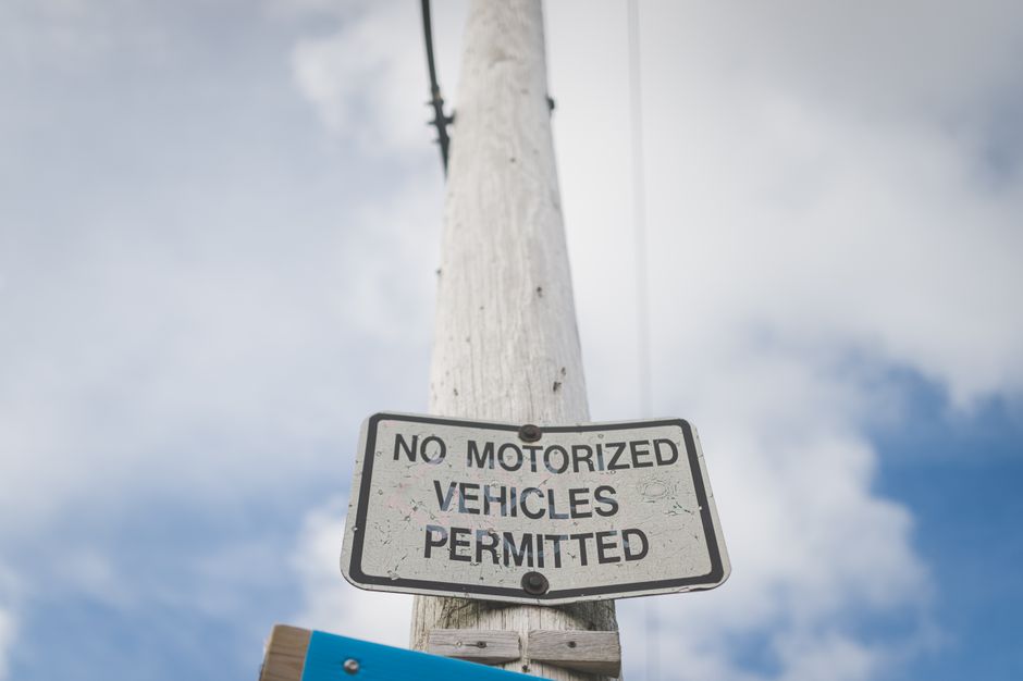 A metal pole mounted against a stone wall exterior of a building, displaying two street parking and loading restrictions signs. The upper sign indicates no parking except by permit from 7 am to 6 pm, Monday to Friday, with a red circle and slash over a 'P' symbol. The lower sign shows designated parking hours from 8 am to 6 pm, Monday to Friday, with a green circle around a 'P' symbol and a hand gesture icon, possibly indicating loading or unloading permitted during these times. Part of a window with a green-tinted glass pane and wooden framing is visible at the top. This signage is relevant for house removals or moving services, as it informs about parking and loading regulations in Old Malden, supporting the logistical planning for a home relocation by Man with Van Old Malden.