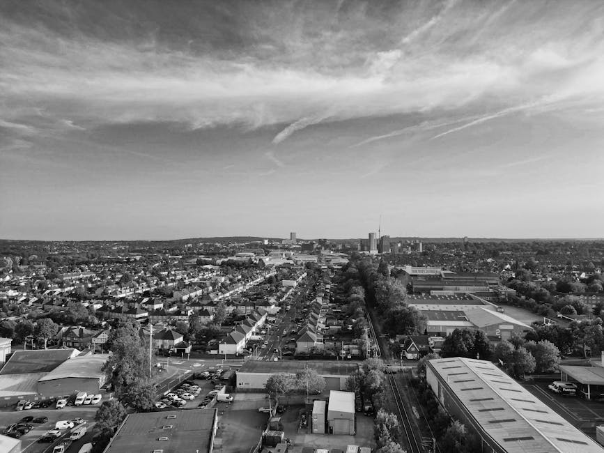 A black and white aerial photograph showing a residential area with a mix of modern apartment buildings, terraced houses, and smaller single-family homes. The image captures a street with parked cars